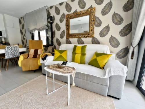 Elegante salón del apartamento en Fuengirola con sofá gris, cojines amarillos, papel tapiz con motivo de hojas y espejo dorado