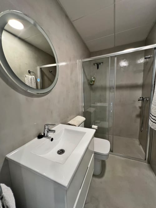 Baño moderno en apartamento de lujo con lavabo blanco, espejo circular y cabina de ducha de cristal en Fuengirola