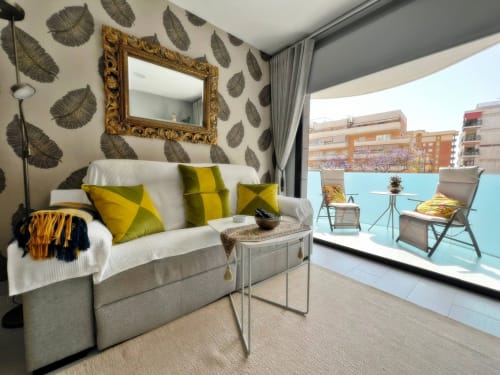 Salón de apartamento de lujo en Fuengirola con sofá moderno, espejo dorado, papel pintado con hojas y balcón terraza.