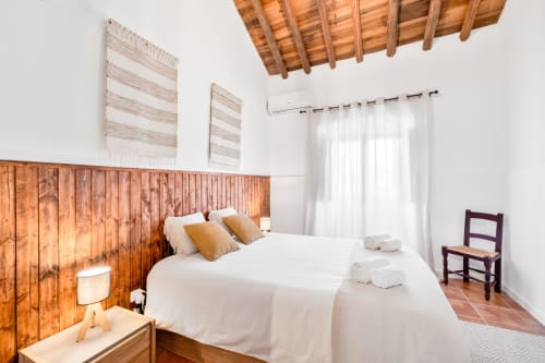 Quarto aconchegante da Casa da Ti Inácia com cabeceira de madeira, roupa de cama branca, teto com vigas aparentes e azulejos de terracota