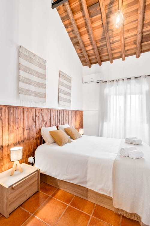 Quarto rústico da Casa da Ti Inácia com vigas de madeira, roupa de cama branca, azulejos de terracota e comodidades modernas