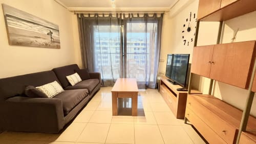 Moderno salón de apartamento con vista al mar en Valencia, sofá oscuro, muebles de madera, TV y balcón frente a la costa