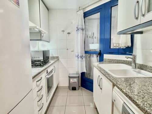 Cocina y baño modernos en apartamento con vistas al mar en Valencia, gabinetes blancos, encimeras de granito, puerta azul y acero inoxidable