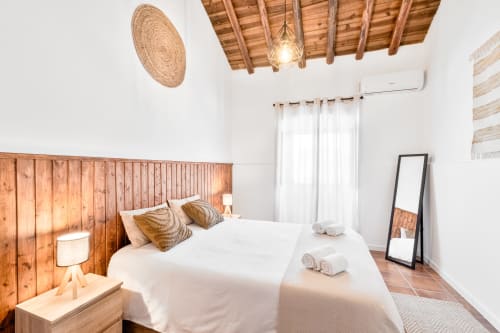 Quarto luminoso da Casa da Ti Inácia com cabeceira de madeira, roupa de cama branca, teto de vigas aparentes e ar-condicionado moderno