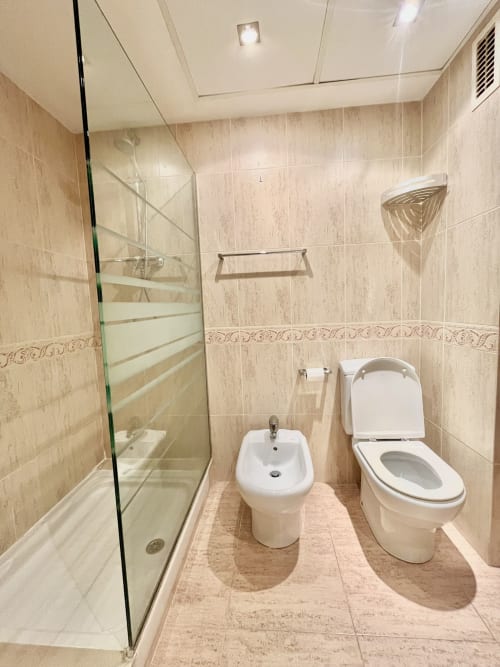 Baño moderno en apartamento con vista al mar en Valencia, ducha de cristal, bidé, inodoro, azulejos beige e iluminación empotrada