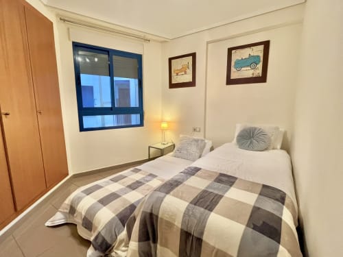 Dormitorio luminoso en apartamento de Valencia con ropa de cama a cuadros, ventana con vista al mar y arte de coches clásicos