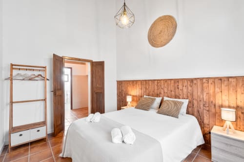 Quarto espaçoso da Casa da Ti Inácia com cabeceira de madeira, roupa de cama branca, toalhas dobradas, guarda-roupa de madeira e parede em tecido