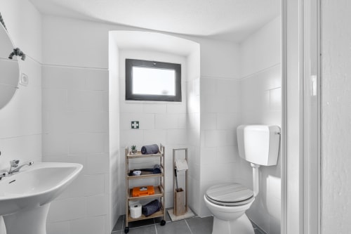 Banheiro moderno e limpo da Casa da Ti Inácia com vaso sanitário, pia, chuveiro e prateleiras de madeira para armazenamento