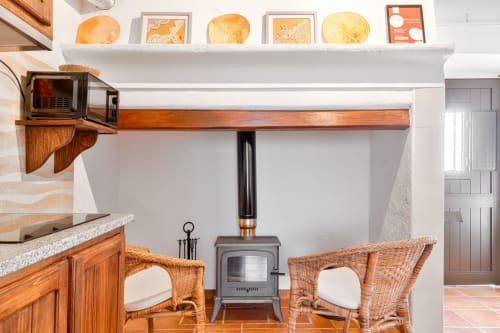 Aconchego na sala da Casa da Ti Inácia com lareira, cadeiras de vime e cozinha decorada com pratos.