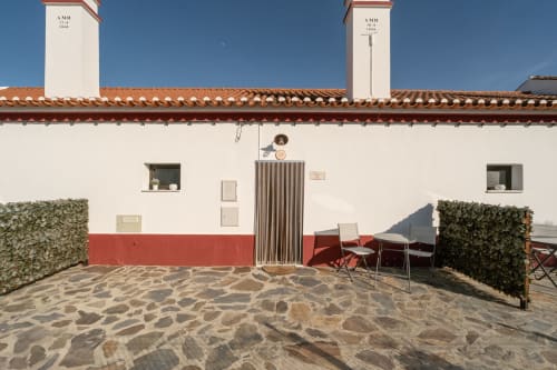 Pátio da Casa da Ti Inácia com piso de pedra, construção vermelha e branca, assentos ao ar livre e chaminés sob céu azul claro
