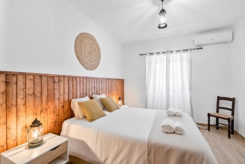 Quarto luminoso da Casa da Ritinha com cabeceira de madeira, roupa de cama branca, almofadas douradas e decoração rústica em Montes Juntos