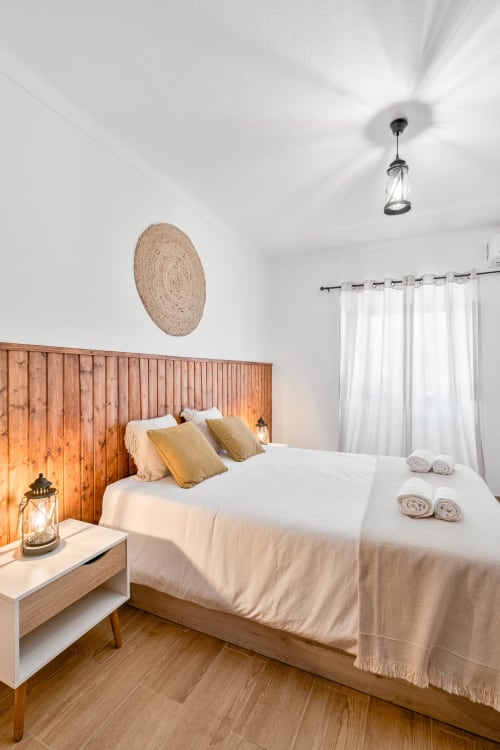Quarto claro na Casa da Ritinha com cabeceira de madeira, roupas de cama brancas, almofadas amarelas e elementos rústicos