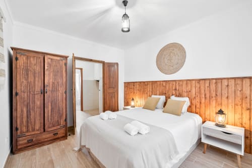 Quarto iluminado da Casa da Ritinha com revestimento em madeira, roupa de cama branca, guarda-roupa rústico e decoração em tecido na parede