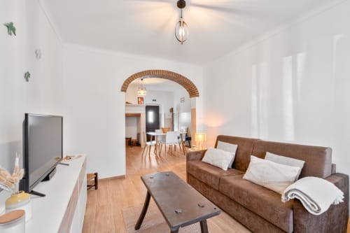 Sala de estar luminosa na Casa da Ritinha com sofá marrom, mesa de jantar, TV e porta em arco de tijolos para a área de refeições
