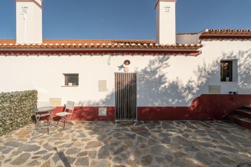 Pátio da Casa da Ritinha em Montes Juntos com fachada vermelha e branca, telhado em barro, piso de pedra, cadeiras vermelhas e vegetação trepadeira