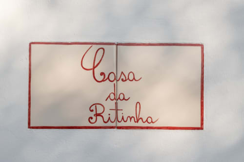 Placa com moldura vermelha com 'Casa da Ritinha' em letras cursivas, placa de boas-vindas no interior do alojamento Montes Juntos
