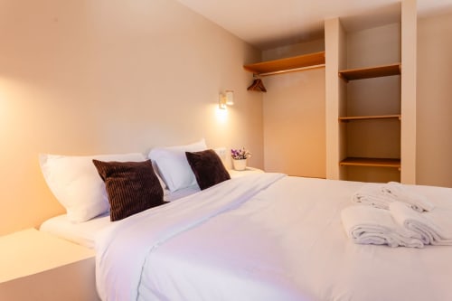 Quarto moderno na FC Housing Company em Abrantes com roupas de cama brancas, almofadas marrons, prateleiras de madeira e iluminação ambiente