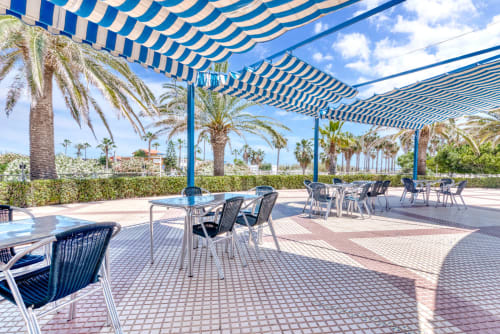 Shaded outdoor dining terrace at Pataconablue Rentals in Valencia with blue and white striped awning, palm trees, and sea