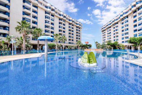 Large outdoor swimming pool at Pataconablue Rentals in Valencia with white apartment buildings, palm trees, and blue