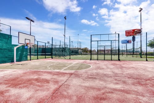 Outdoor sports courts at Pataconablue Rentals in Valencia with basketball hoop, red flooring, green fencing, and blue sky.