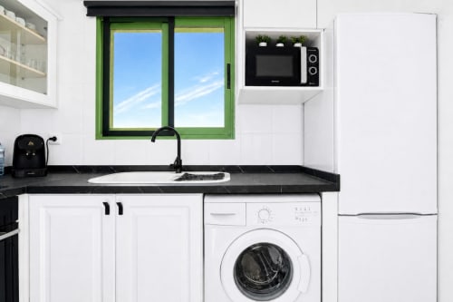 Modern kitchen with white cabinets, black countertop, washing machine, green-framed sea view window at Valencia apartment