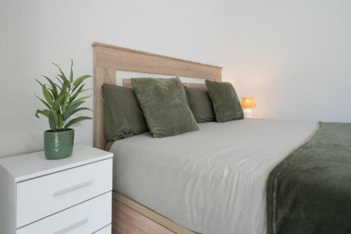 Dormitorio moderno en el hotel Studios Sanchis de Alicante con cama de madera, almohadas verdes, ropa blanca y planta en maceta