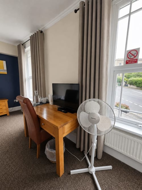 The Cornerhouse Triple Room workspace with wooden desk, brown leather chair, TV, white standing fan, and beige curtains
