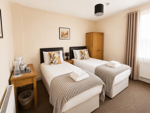 Twin beds with grey throws in a beige bedroom at The Cornerhouse hotel, Somerset, with wooden wardrobe and side table.