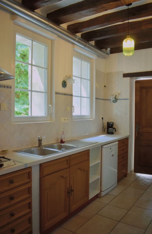 Cuisine moderne avec placards en bois, évier en acier inoxydable et fenêtres donnant sur les jardins du gîte Le Moulin Garnier