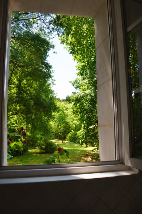 Vue depuis la fenêtre ouverte du jardin verdoyant et du paysage bordé d'arbres du gîte Le Moulin Garnier à Vernou-sur-Brenne