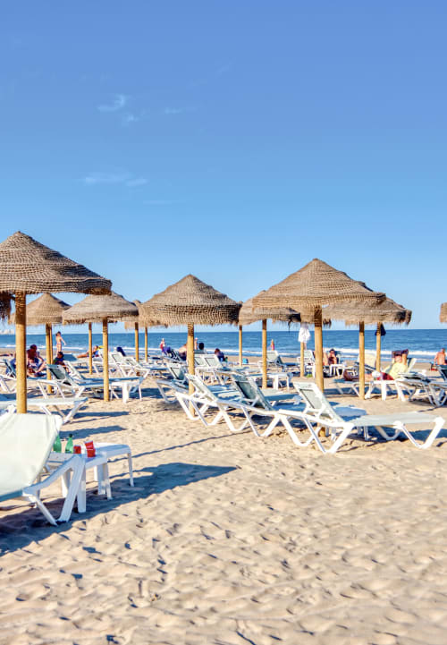 Playa de arena con sombrillas de paja y tumbonas blancas con vistas al mar Mediterráneo en Valencia, España