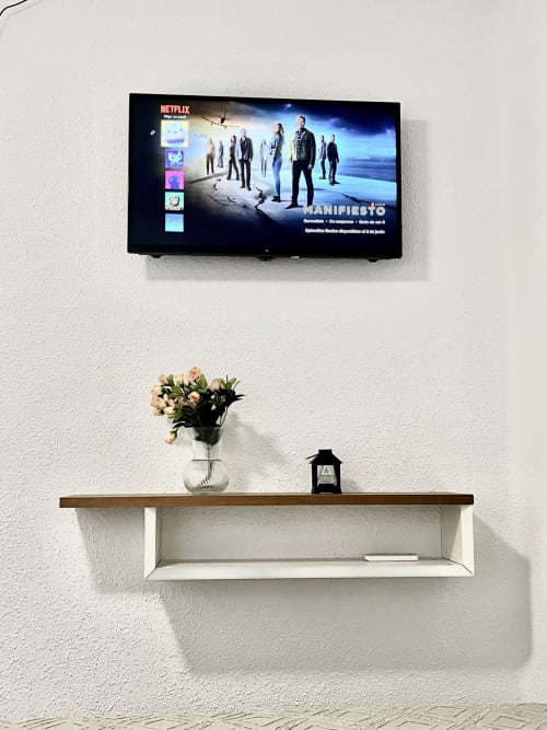 Sala de estar moderna con televisor montado en pared mostrando Netflix, estantería de madera con flores y accesorios decorativos, pared blanca