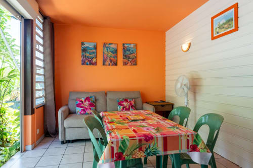 Chambre lumineuse en Guadeloupe aux murs orange, décoration tropicale, chaises vertes, literie colorée et accès à la terrasse