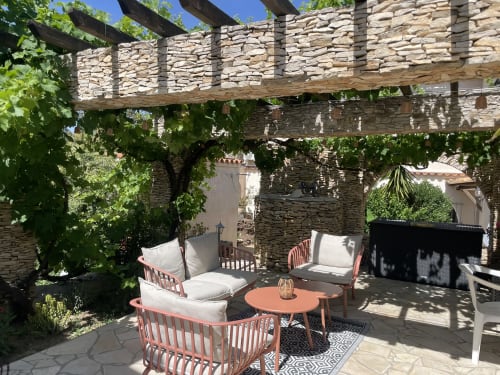 Terrasse ombragée du gîte La Closerie à La Redorte avec chaises corail, table ronde et verdure luxuriante sous pergola en pierre