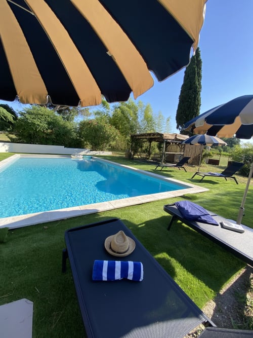 Piscine avec transats, parasols rayés et jardins luxuriants au gîte Le Domaine les Olivoux à Pomport, France