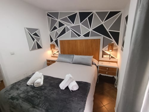 Dormitorio moderno en villa Las Mellis de Antequera con cabecero de madera, almohadas grises, toallas blancas y arte geométrico en la pared