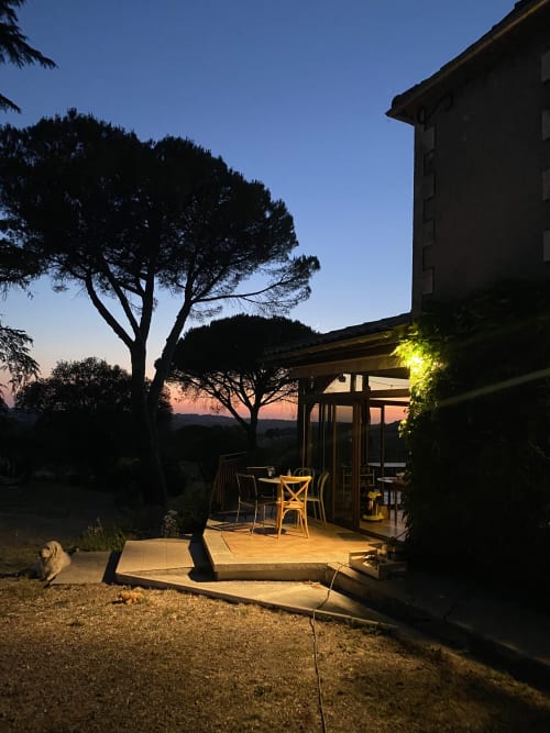 Terrasse au crépuscule avec salle à manger illuminée, arbre et chien au gîte Le Domaine les Olivoux à Pomport
