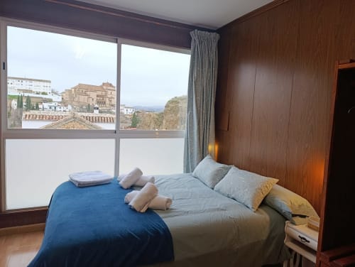 Dormitorio moderno con ropa de cama azul y gris, paredes de madera y grandes ventanales con vistas al casco antiguo histórico de Antequera