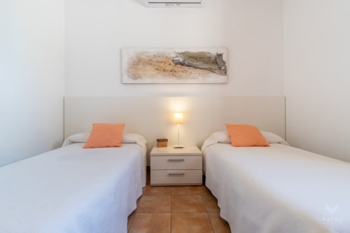 Dos camas gemelas con almohadas naranjas en dormitorio de villa Artá, luz natural de lámpara de mesita, decoración minimalista
