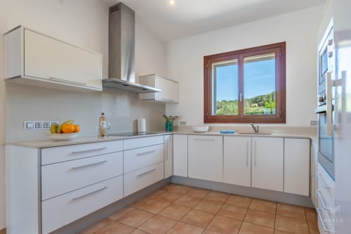 Cocina blanca moderna con azulejos de terracota, campana de acero inoxidable y ventana con marco rojo con vistas a las montañas de Artá