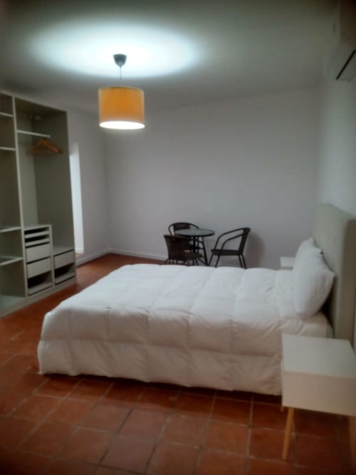 Quarto luminoso na pousada Casa Augusta com cama branca, luminária de pendente laranja, piso de madeira, guarda-roupa e área de estar