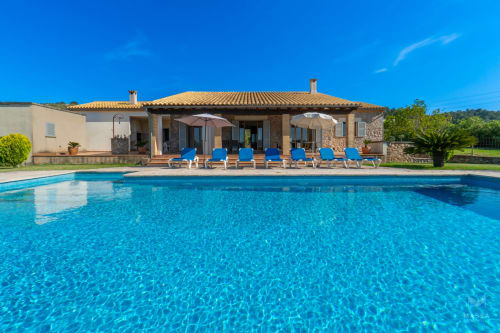 Villa de lujo en Artà con piscina cristalina, tumbonas azules y casa con tejado de barro bajo cielo despejado