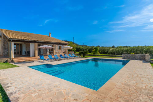Villa de lujo en Artà con piscina resplandeciente, tumbonas azules y vistas a viñedos bajo cielo despejado
