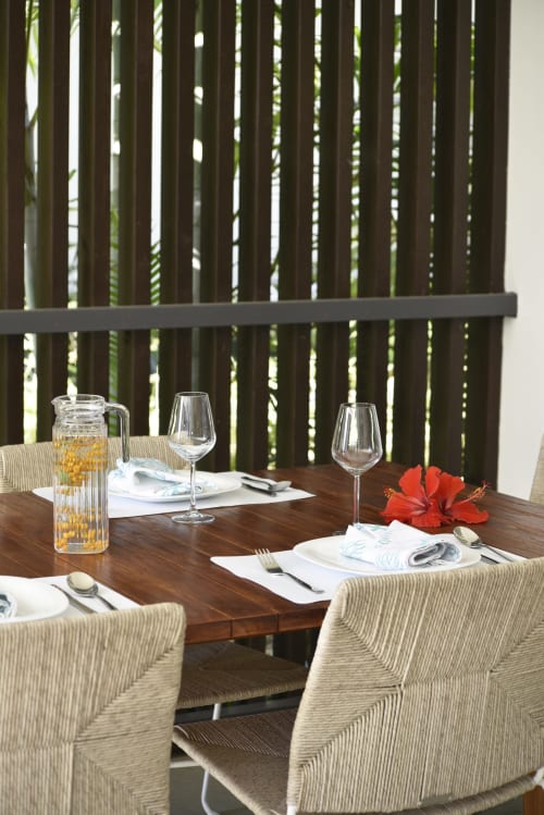 Outdoor dining area at O'Biches apartment with wooden table, woven chairs, place settings, red hibiscus flower, and slatted