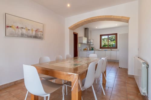 Comedor moderno con mesa de madera y sillas blancas que se abre a cocina con puerta arqueada en propiedad de Artà