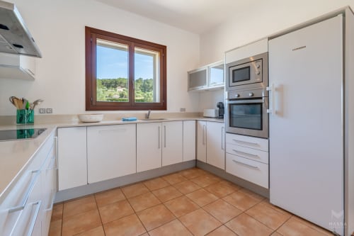 Cocina blanca moderna con azulejos de terracota, ventana de marco de madera y electrodomésticos de acero inoxidable en Artà, España