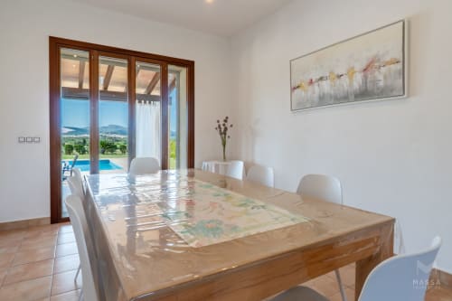 Comedor moderno con mesa de madera, sillas blancas y vistas panorámicas de la campiña de Artà y piscina
