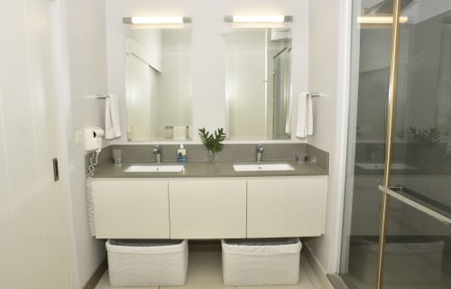 Modern bathroom vanity with dual sinks, grey countertop, white cabinetry, mirrors with lighting, and shower at Pointe d'Esny