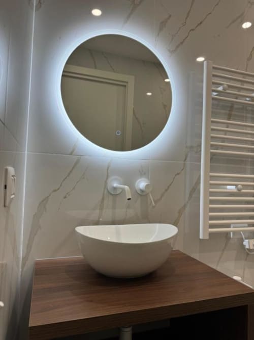 Moderno mobile bagno con lavabo in ceramica bianca, specchio circolare illuminato e rivestimento in marmo presso Maison ì Room in