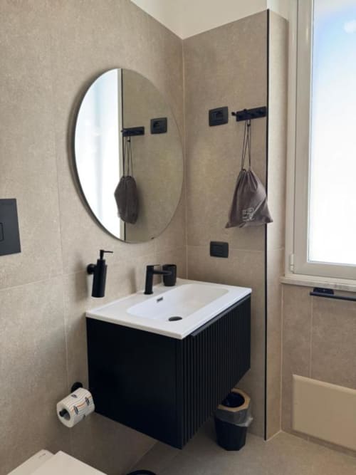 Moderno bagno con specchio rotondo, vanità nera, lavabo bianco e pareti piastrellate neutre presso Maison ì Room a Reggio Calabria